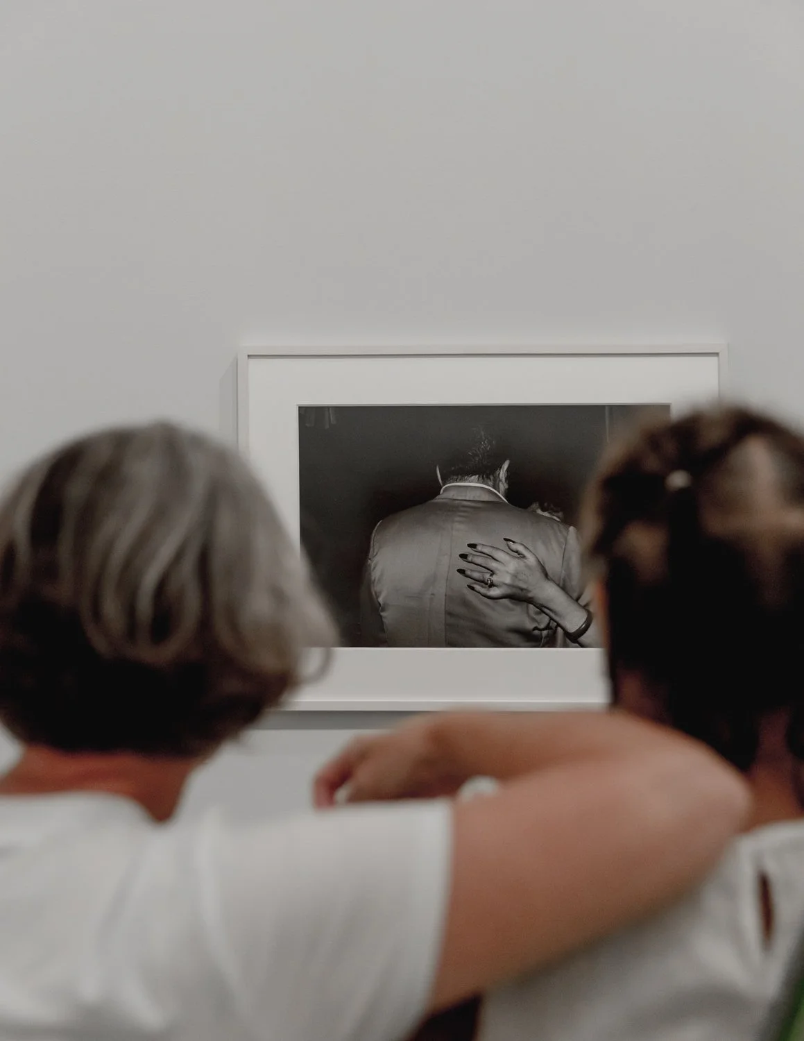 Two women sitting on a bench, looking at a black and white photograph of a person from behind, with the person's arm around them.
