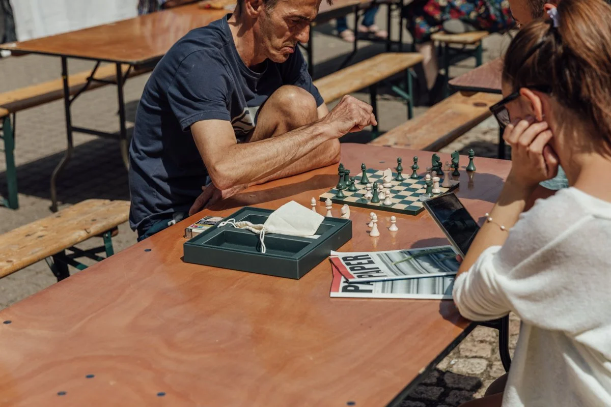 An older man and a young woman are playing chess outdoors at a long wooden table amid empty benches; the man is making a move, while the woman is reading or studying a magazine and holding her head in her hand.