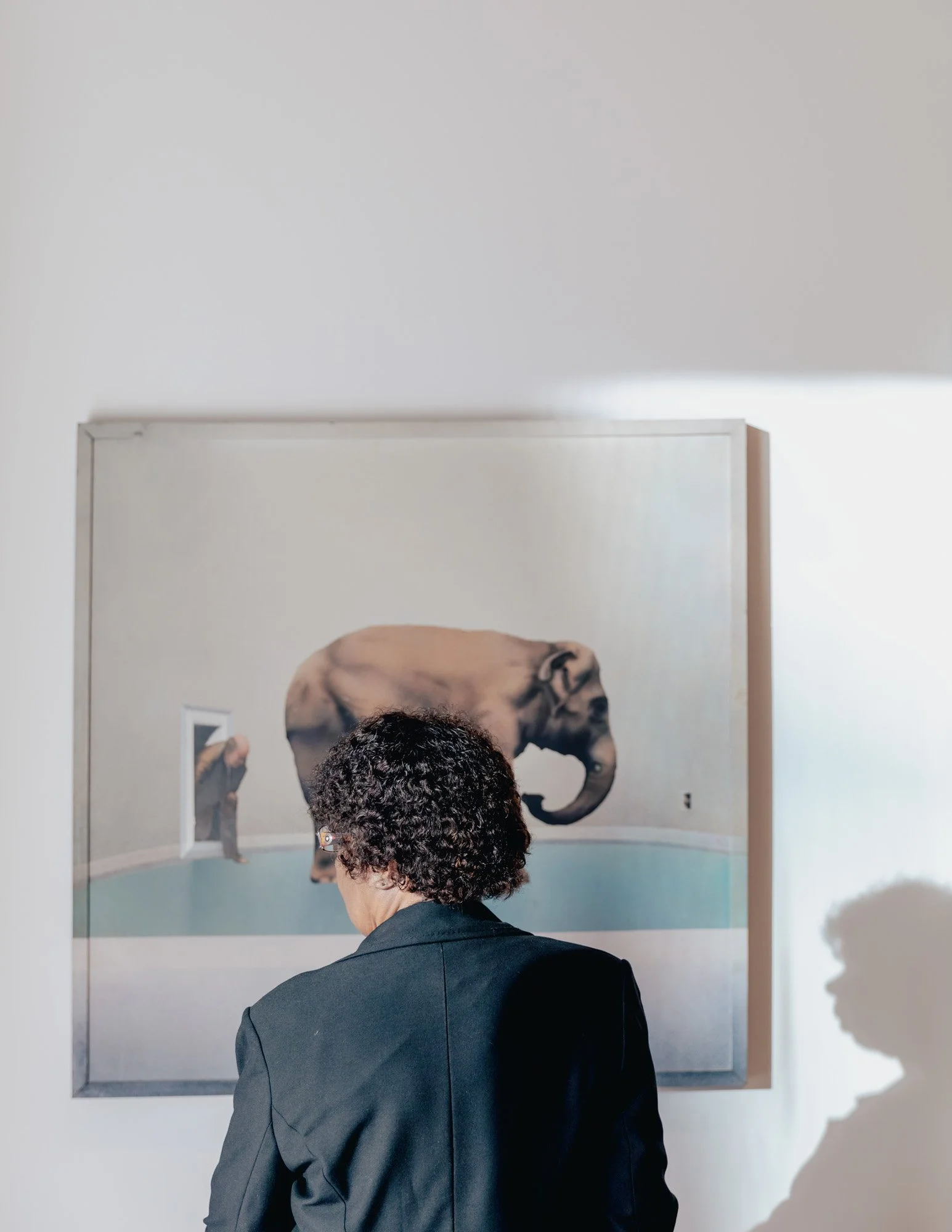 Person with curly hair in a black blazer viewing a surrealist artwork featuring a giant elephant with a tiny human figure in a gallery.