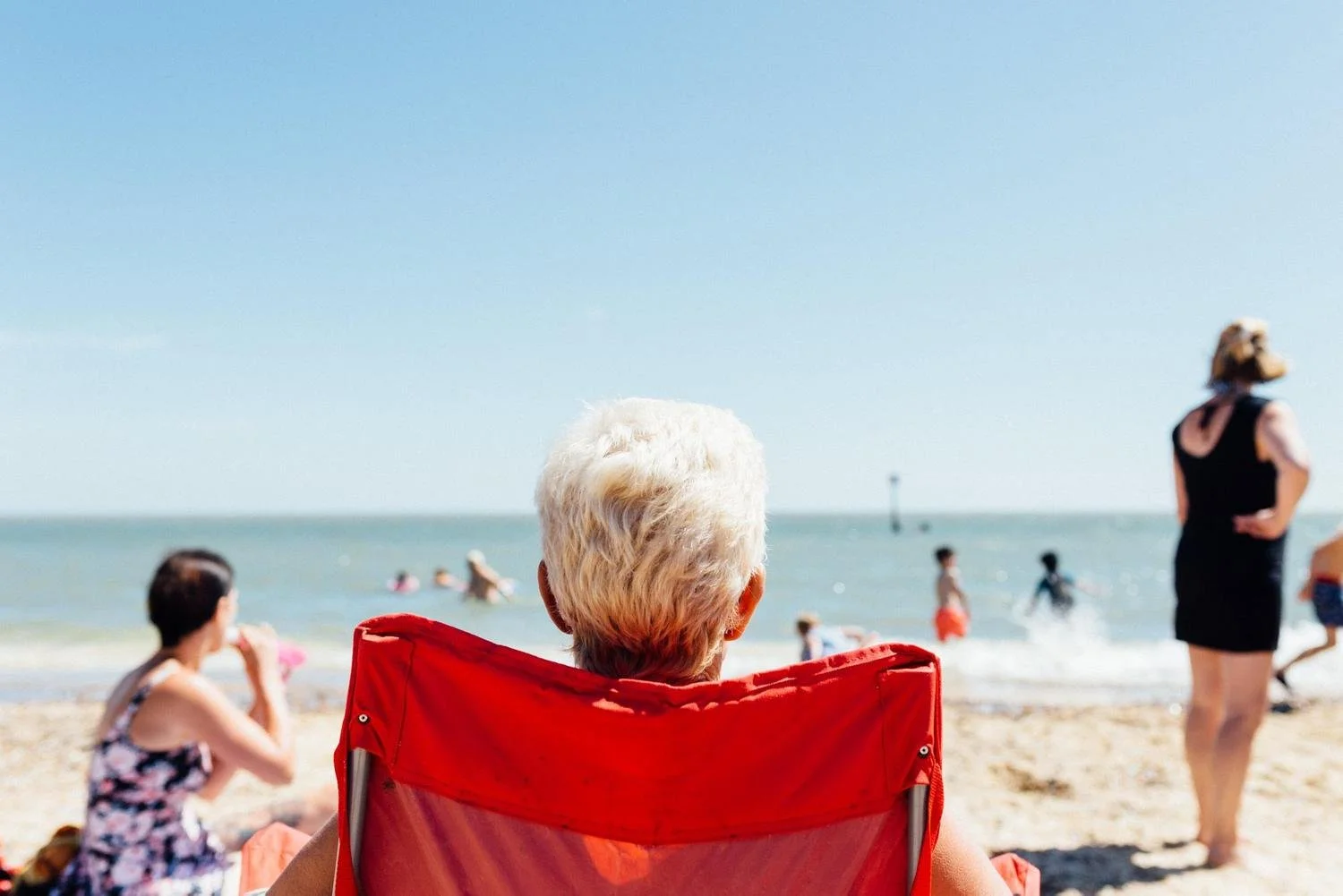 Older woman with white hair sitting in a red beach chair on the sand, facing the ocean, with people swimming and walking along the shoreline on a sunny day.