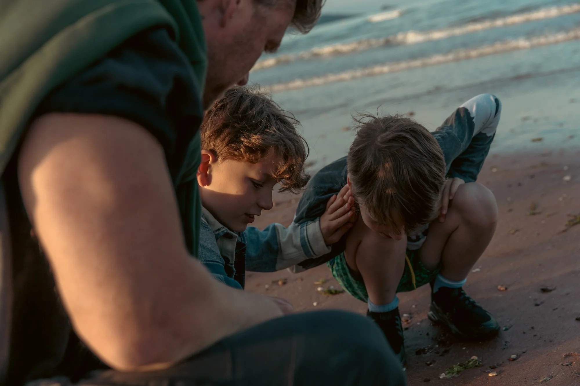 A man with two young boys on a beach, helping one boy to sit down as they play near the water.