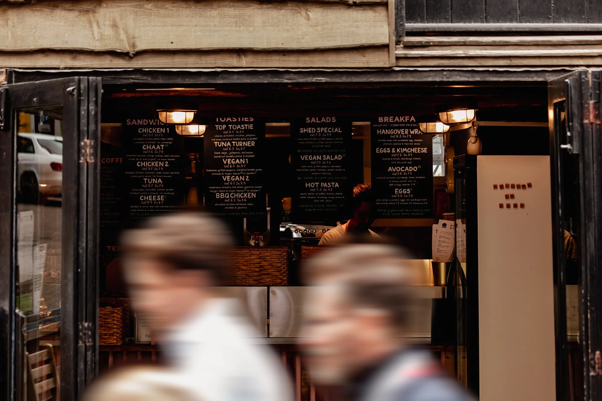A blurry photo of people walking past a small restaurant or cafe with a black chalkboard menu inside. The menu features various food options like sandwiches, salads, breakfast items, and beverages. The interior has warm lighting, and there is a visib