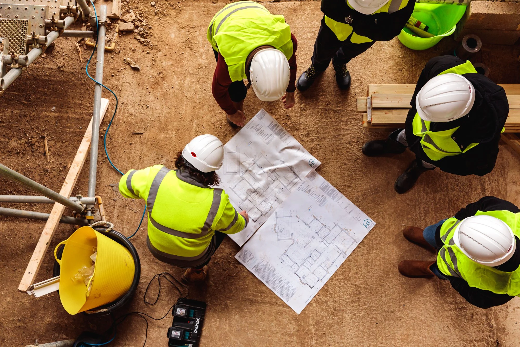 Construction workers wearing safety helmets and high-visibility jackets gathered around blueprints on a dirt floor at a construction site.