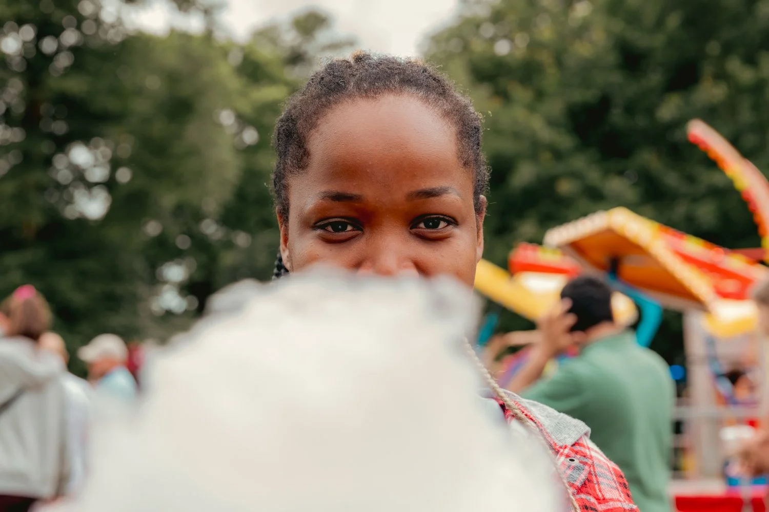 A young Black woman with braided hair holding cotton candy at an outdoor fair, with amusement rides and people in the background.