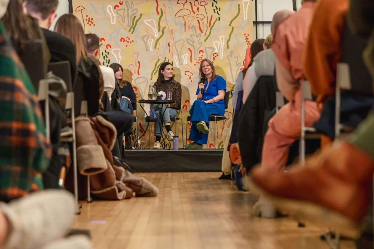 A panel discussion with two women speakers on stage in front of an audience. The woman on the right is wearing a blue dress and holding a microphone, smiling. The woman on the left is wearing a patterned shirt and jeans, attentively listening. The ba