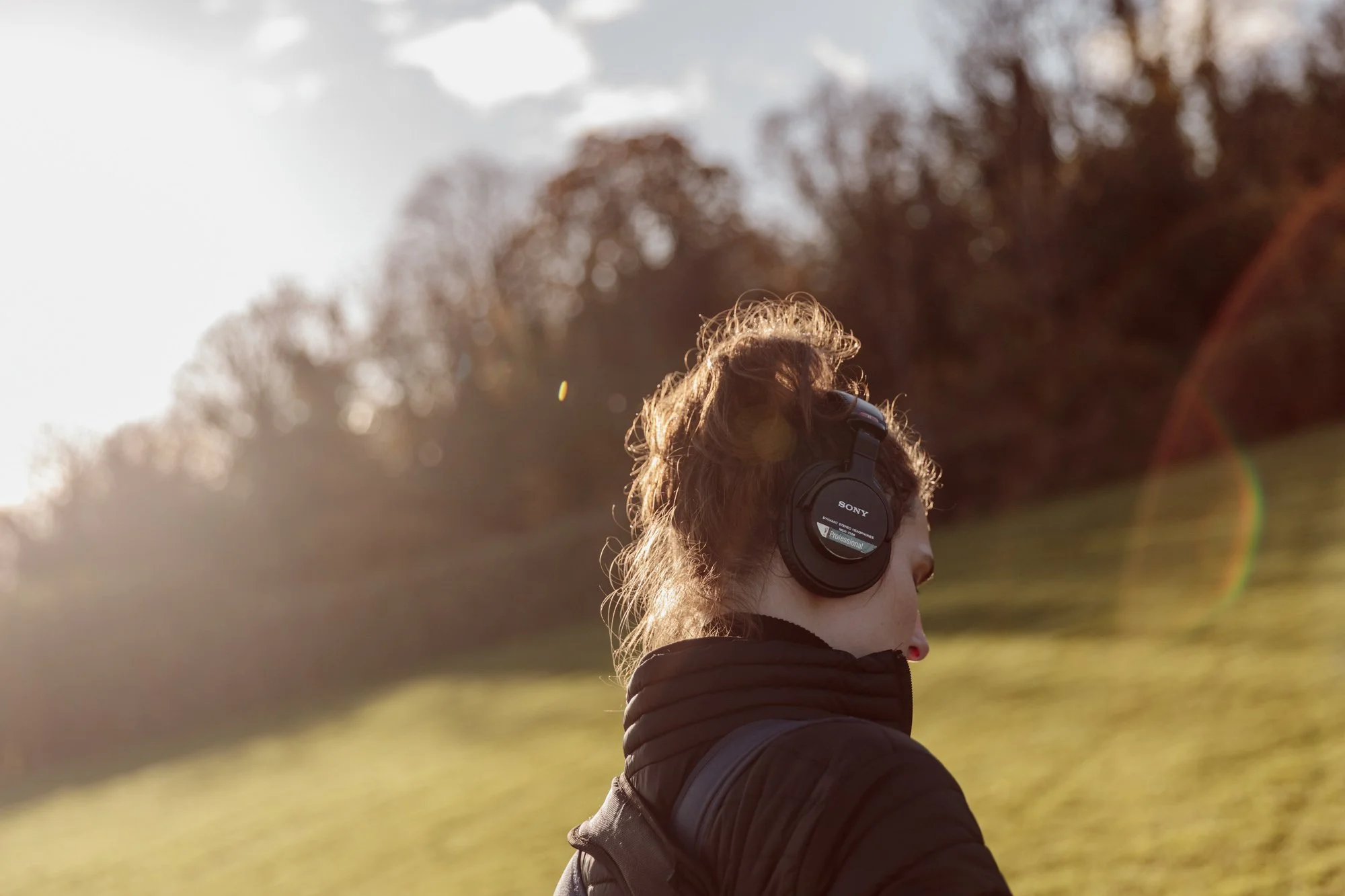 A person with curly hair wearing headphones and a black jacket outdoors on a sunny day, with a blurred background of trees and grass.