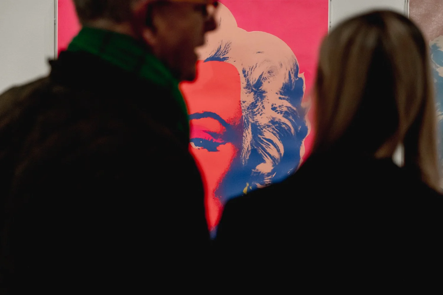Two people observing a pop art portrait of Marilyn Monroe on a pink background, with the portrait rendered in blue and white tones.