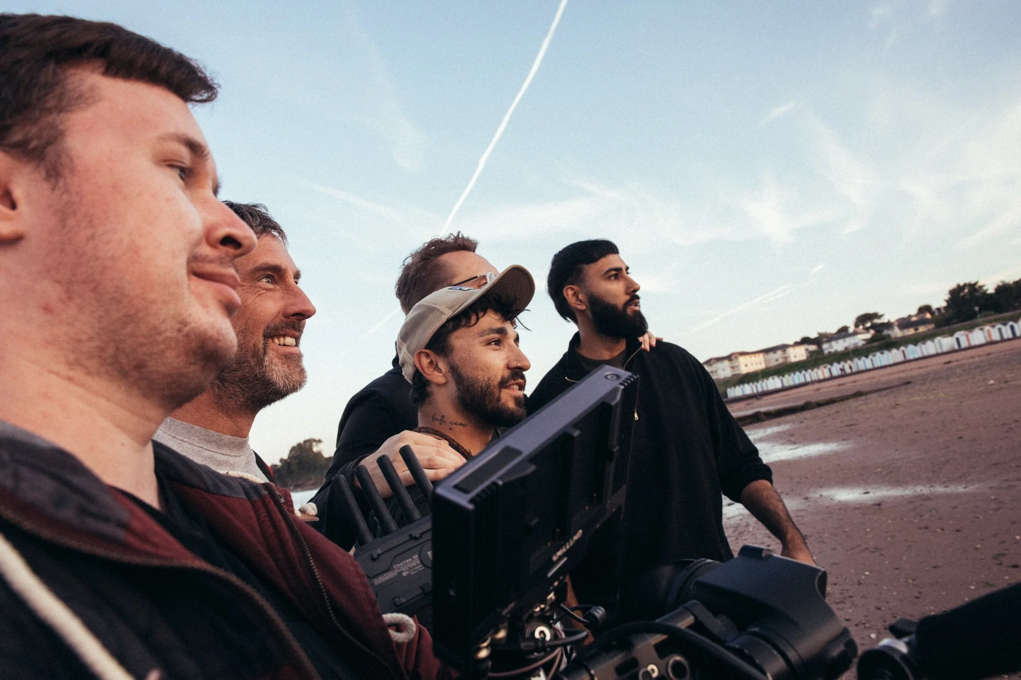 A group of five men standing on a beach, filming a scene with professional camera equipment during sunset or sunrise.