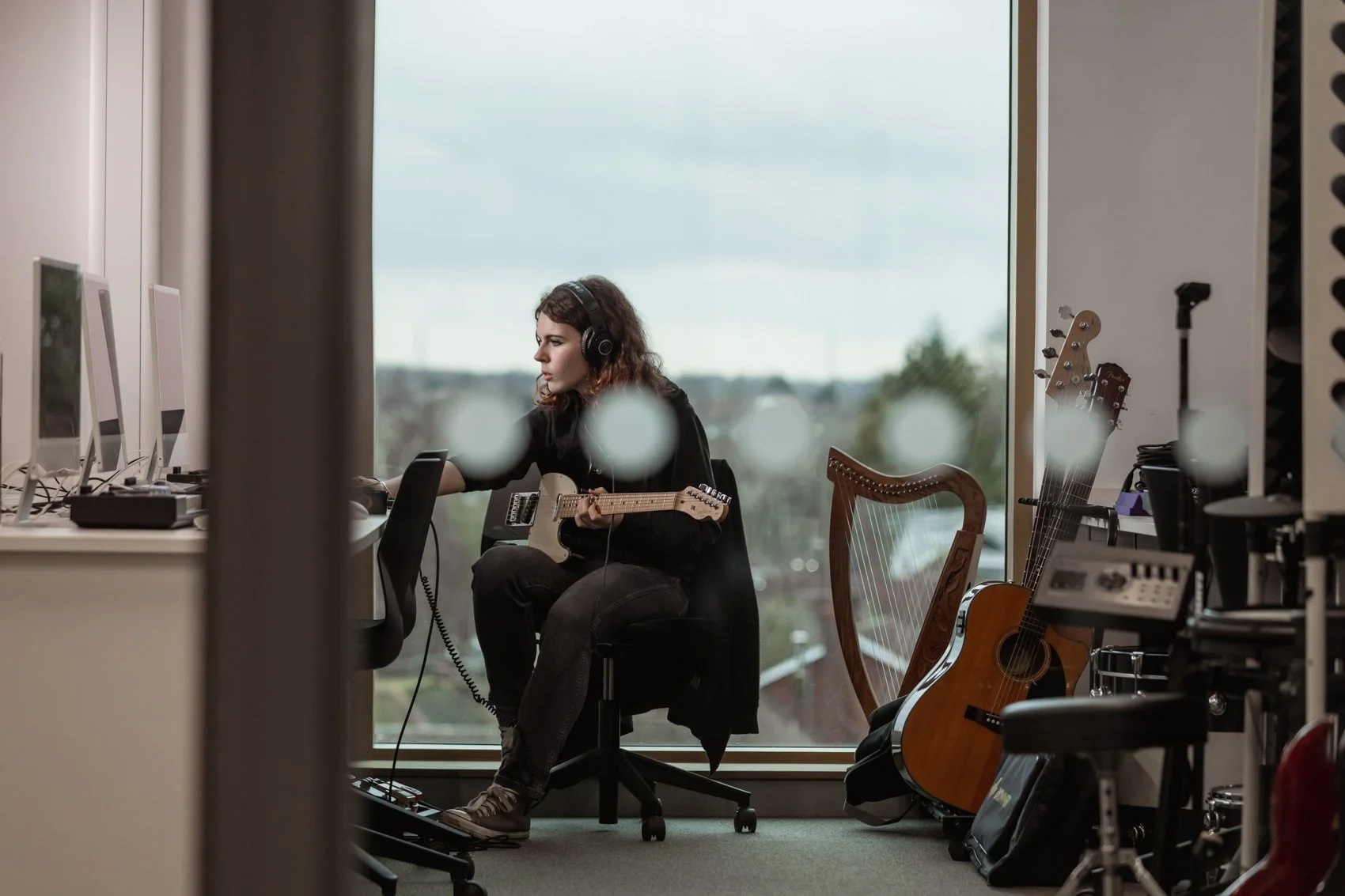 A woman with brown curly hair wearing headphones sits at a desk with computer monitors, playing an electric guitar. There are other guitars, a harp, and music equipment nearby in a music studio with large windows.