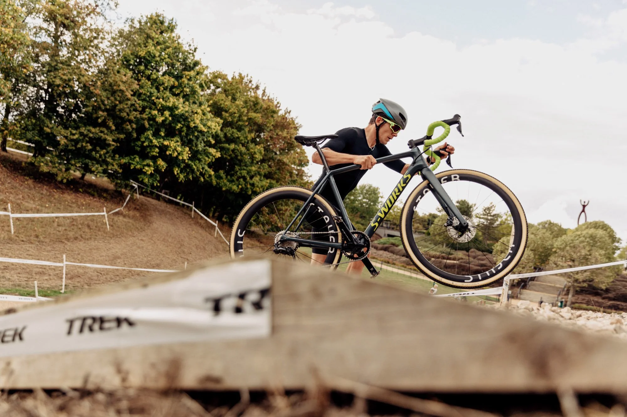 A cyclist carrying a black and green Trek Sworks bicycle on his shoulder during a race or training session outdoors surrounded by trees and a partly cloudy sky.