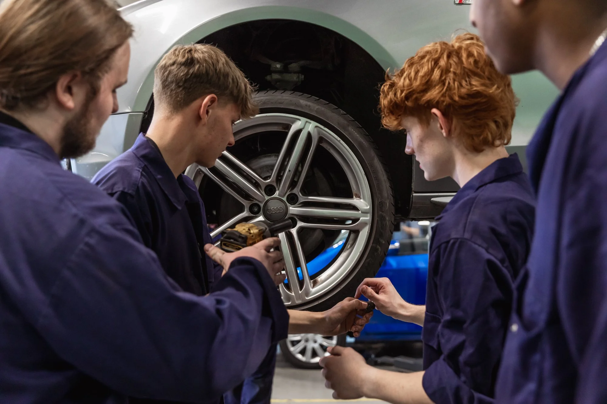 Four young people in mechanic uniforms working on a car in a workshop, with a focus on a wheel and a person using a power tool.