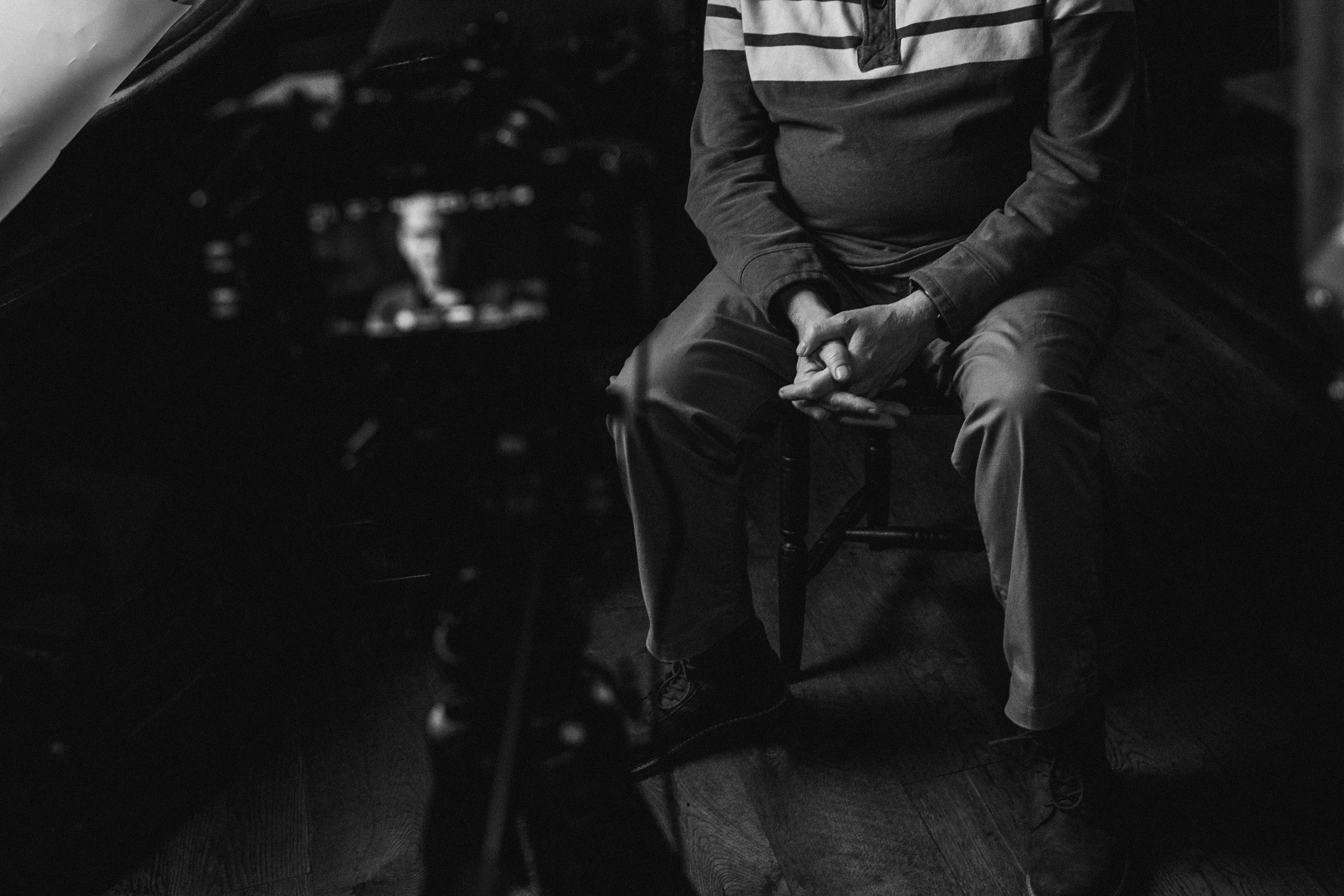 A person sitting on a chair with their hands clasped, wearing a long-sleeve shirt with stripes on the chest, photographed in black and white.