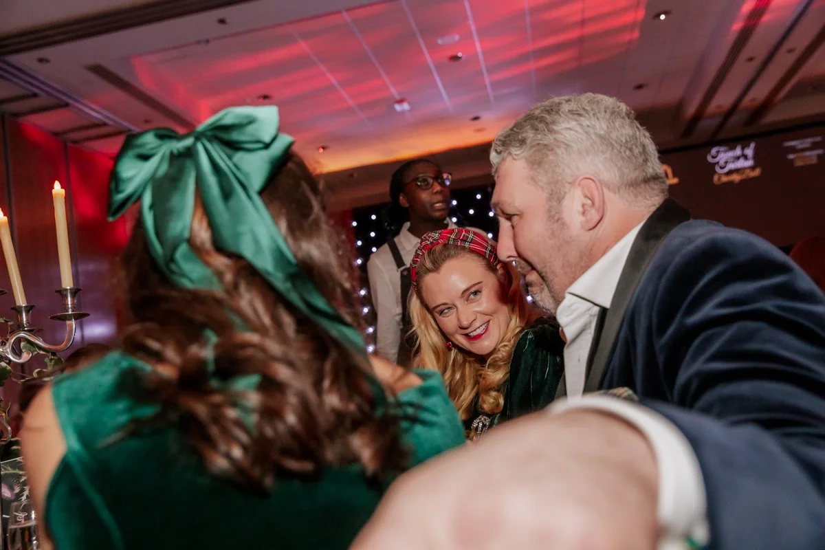 People in festive attire at a party, including a woman with a green bow and dress, a smiling woman with a red headband, and a man in a tuxedo, engaging in conversation.