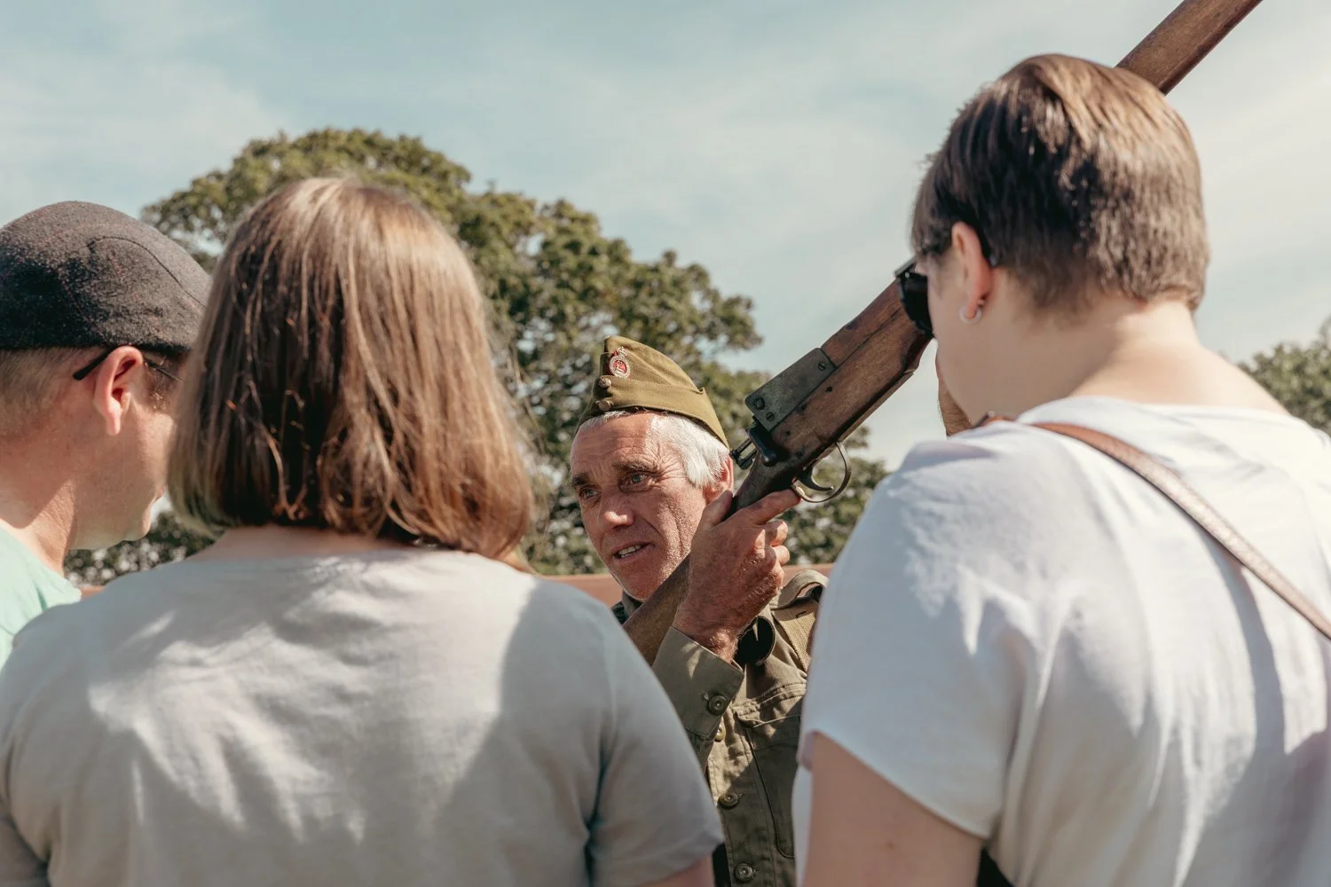 A group of young people listening to a man in a military uniform holding a rifle during an outdoor event, with trees and a cloudy sky in the background.