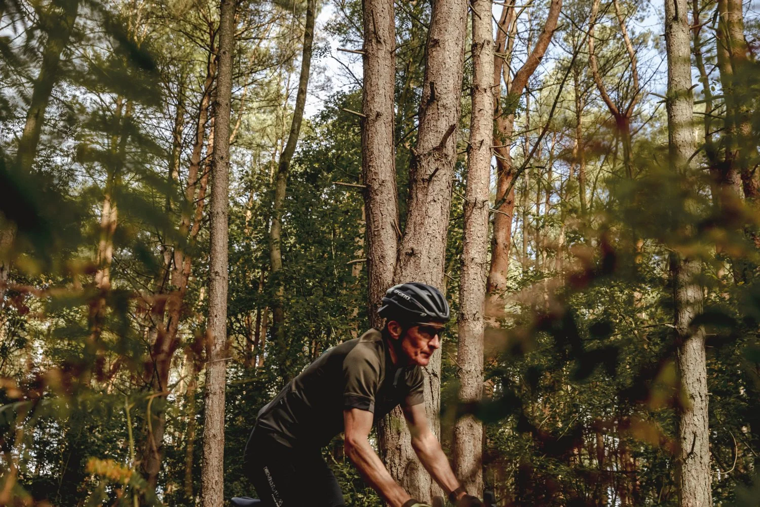 A man wearing a helmet, sunglasses, and a grey cycling jersey riding a bicycle through a forest with tall trees and green foliage.