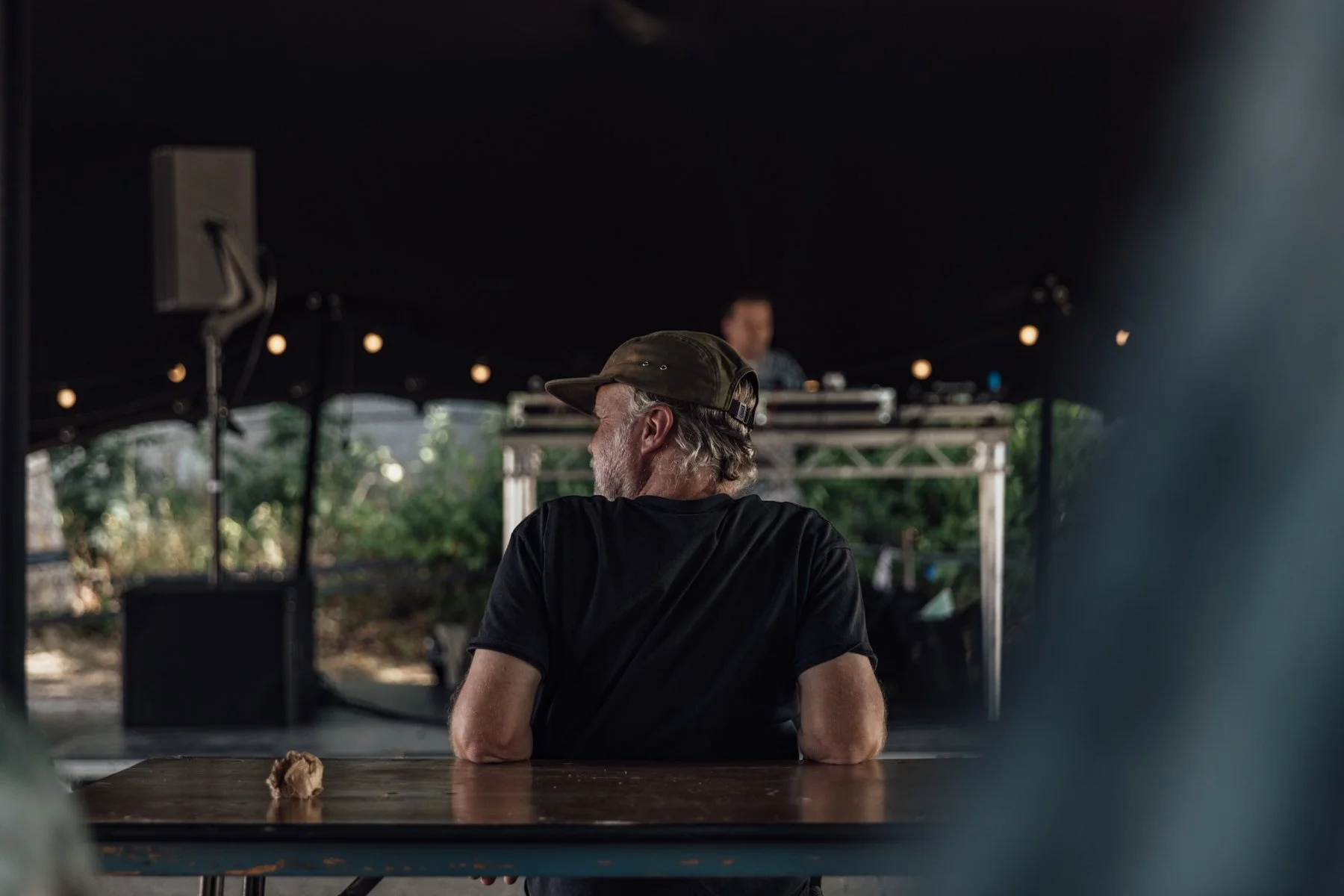 A man with gray hair and beard, wearing a black shirt and a cap, sitting at a wooden table with his back to the camera, in front of a DJ setup with a person in the background, under a dark canopy at an outdoor event during daytime.