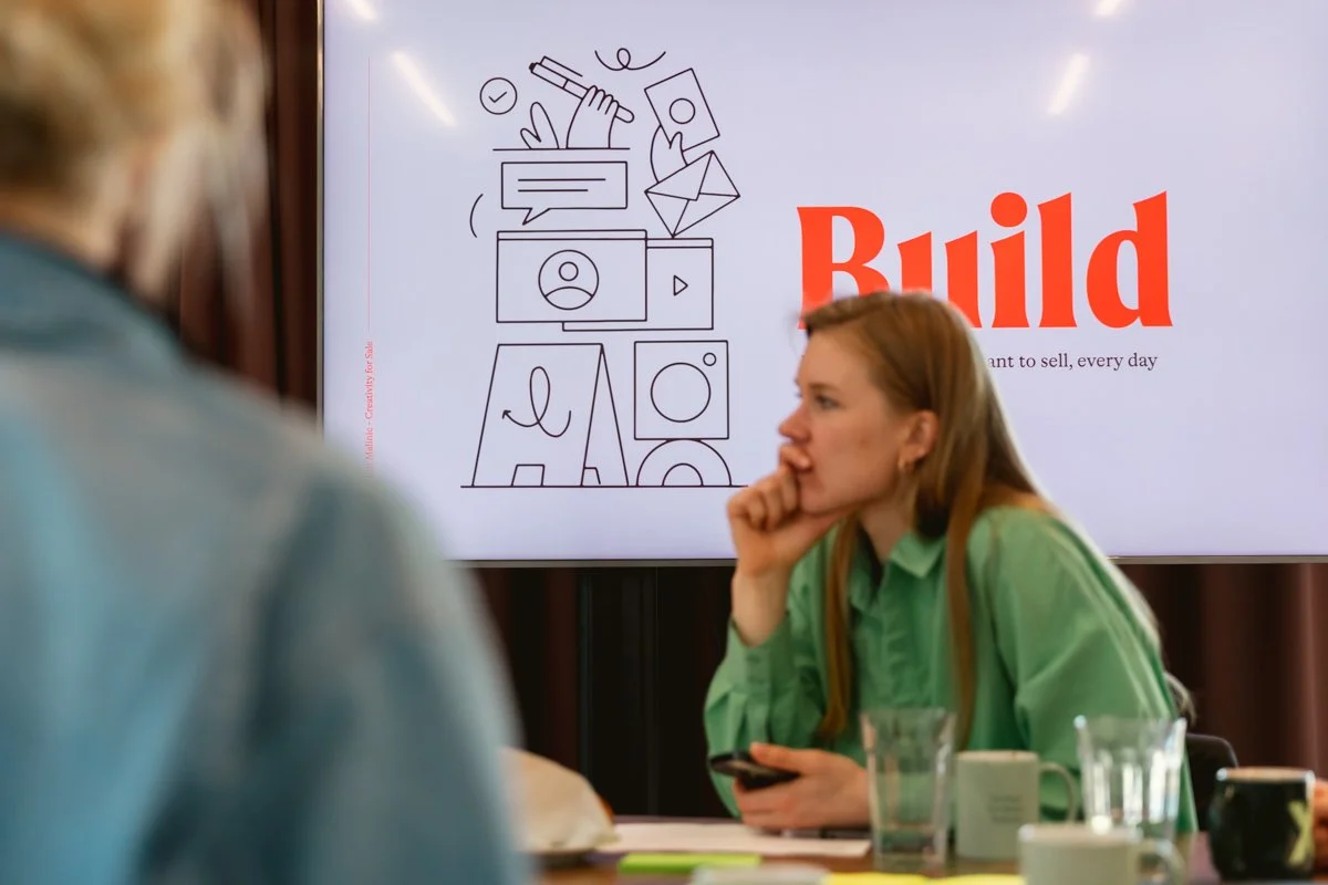 A woman in a green shirt sits at a table, looking thoughtfully at a presentation screen that displays the word 'Build' in large red letters and various outlined icons related to communication and design.
