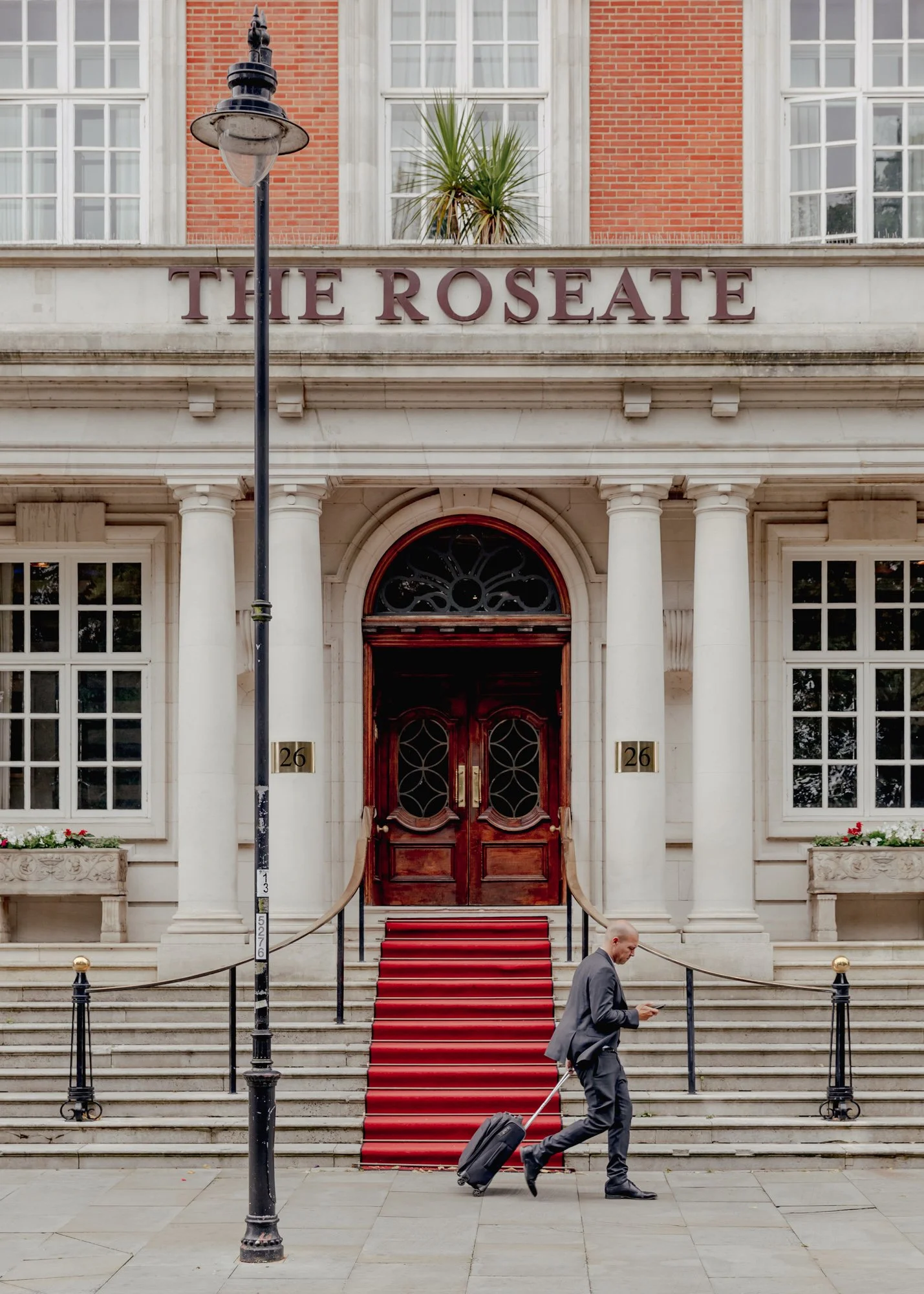 A man in a business suit with a rolling suitcase walking past the entrance of The Roseate hotel, which has white marble steps, a red carpet, large wooden double doors, and a sign with the hotel name above the entrance.