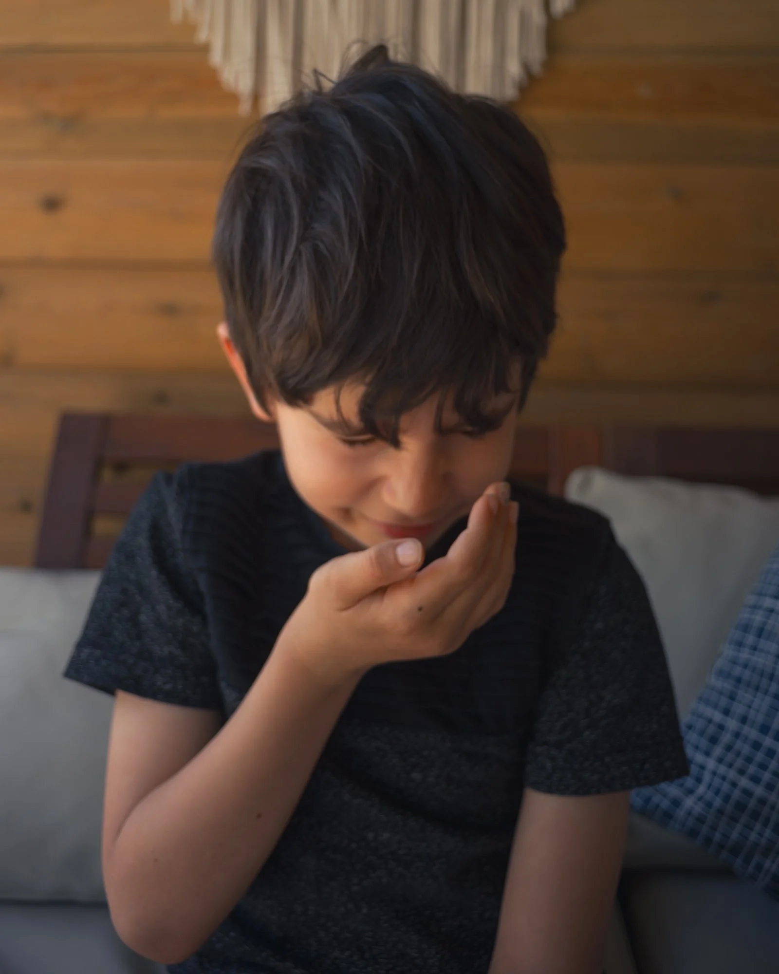 Young boy with dark hair and a black shirt smelling his hand while sitting indoors against wood-paneled wall.
