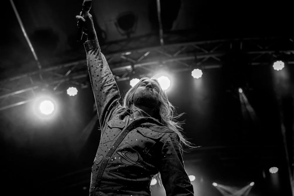 A male singer performing live on stage, microphone in hand, with stage lights shining from behind, black and white photo.