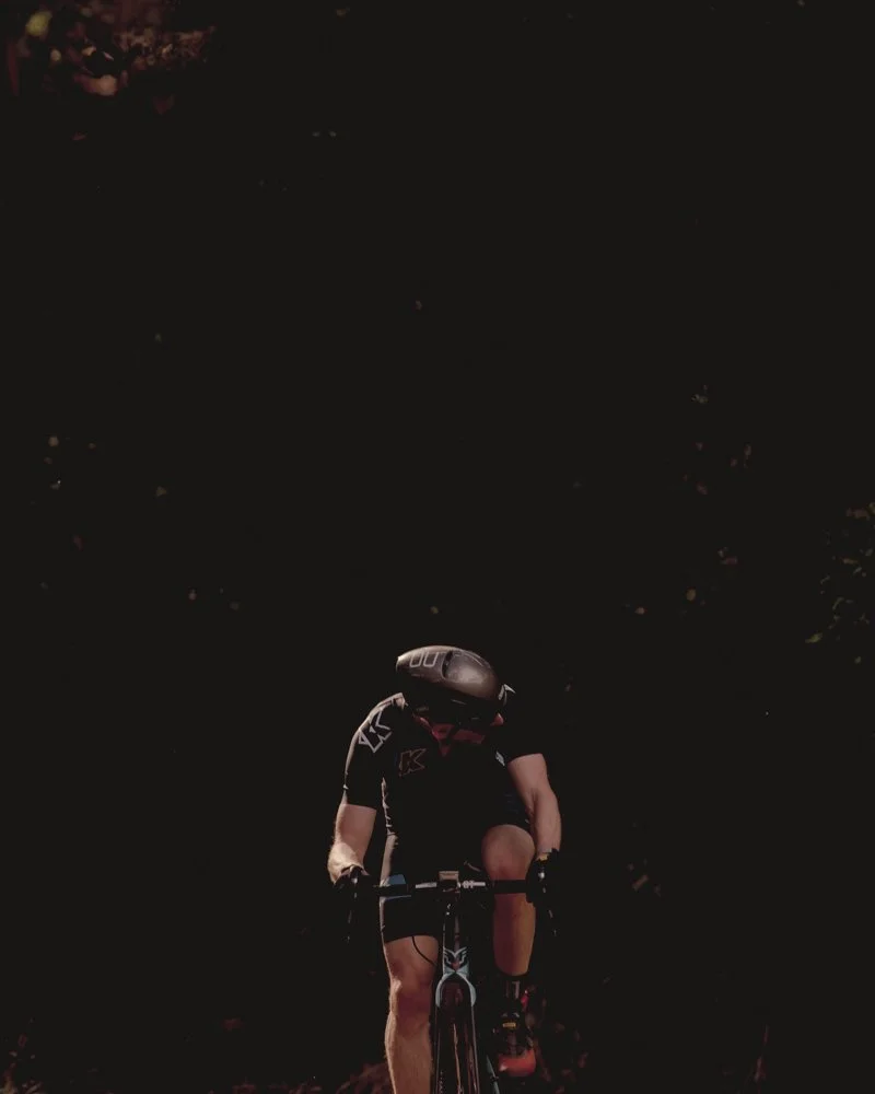 Person riding a mountain bike at night, wearing a helmet and sports clothing, with trees and dark sky in the background.
