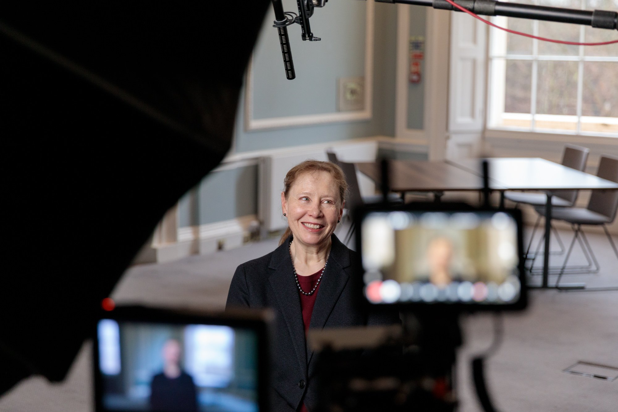 A woman with short reddish hair smiling during an interview, sitting in a well-lit room with large windows, for a video recording with a camera and microphone visible in the foreground.