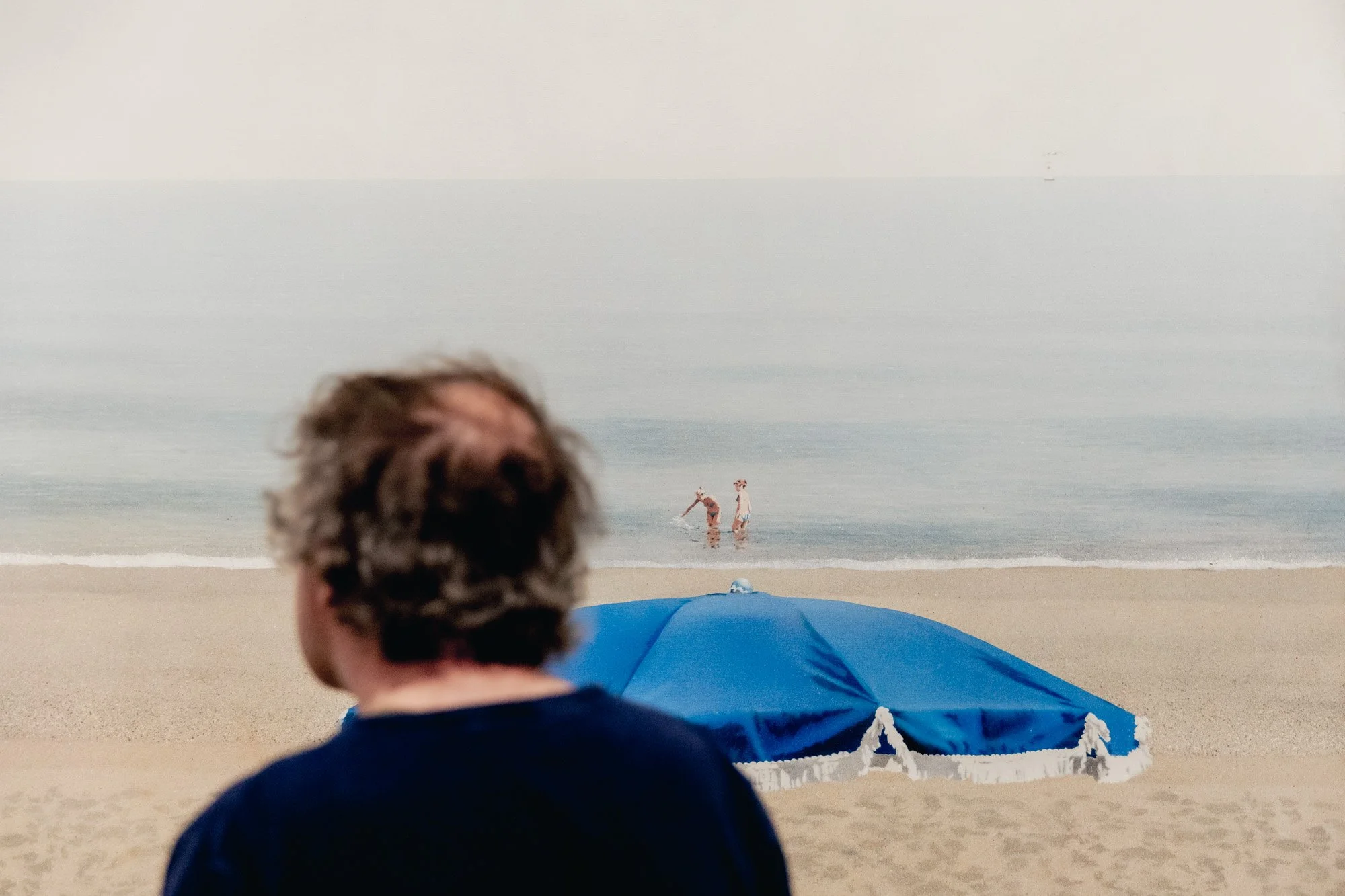A person with curly hair facing away from the camera, watching two people in the ocean with a blue beach umbrella in the foreground.
