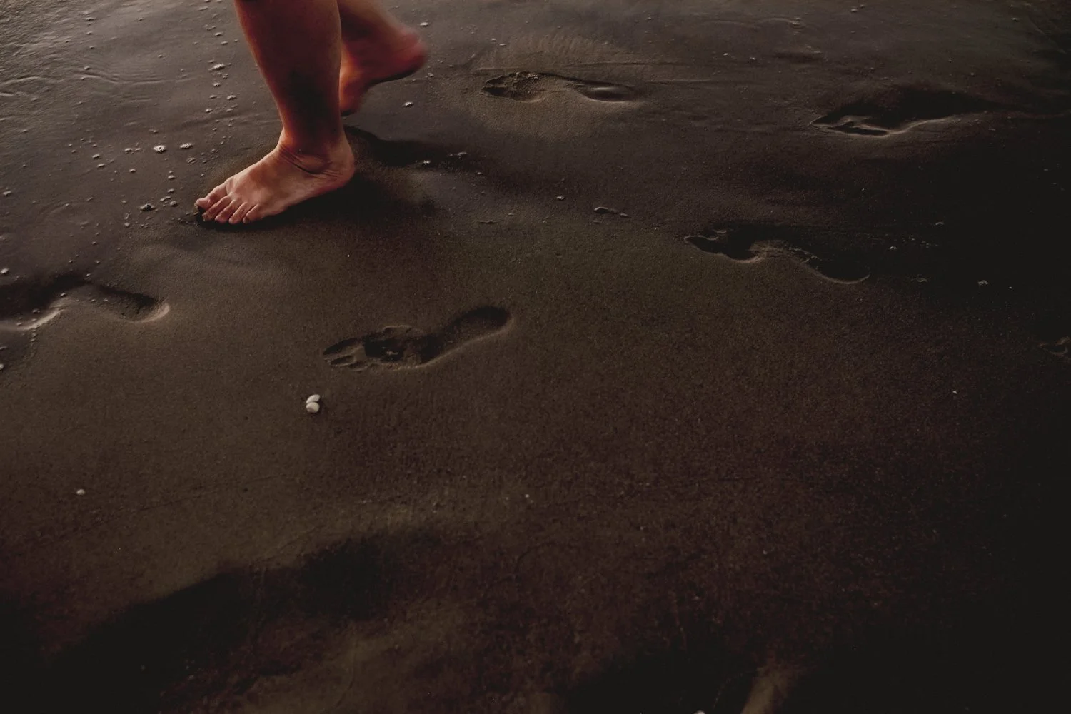 Footprints in dark sand near the shoreline of a beach with a person's bare feet visible in the upper part of the image.