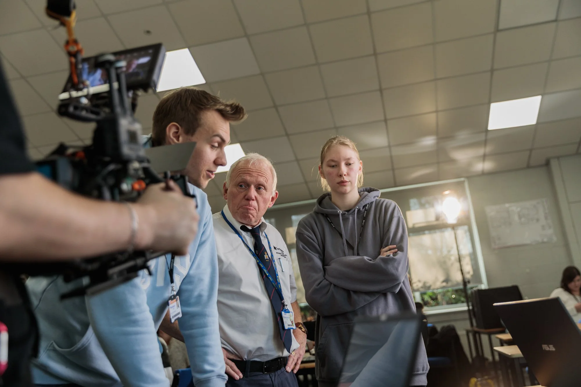 A group of four people, three men and one young woman, appears to be engaged in a discussion or presentation in a classroom or office setting. One man is holding a camera, and all are looking at a laptop.