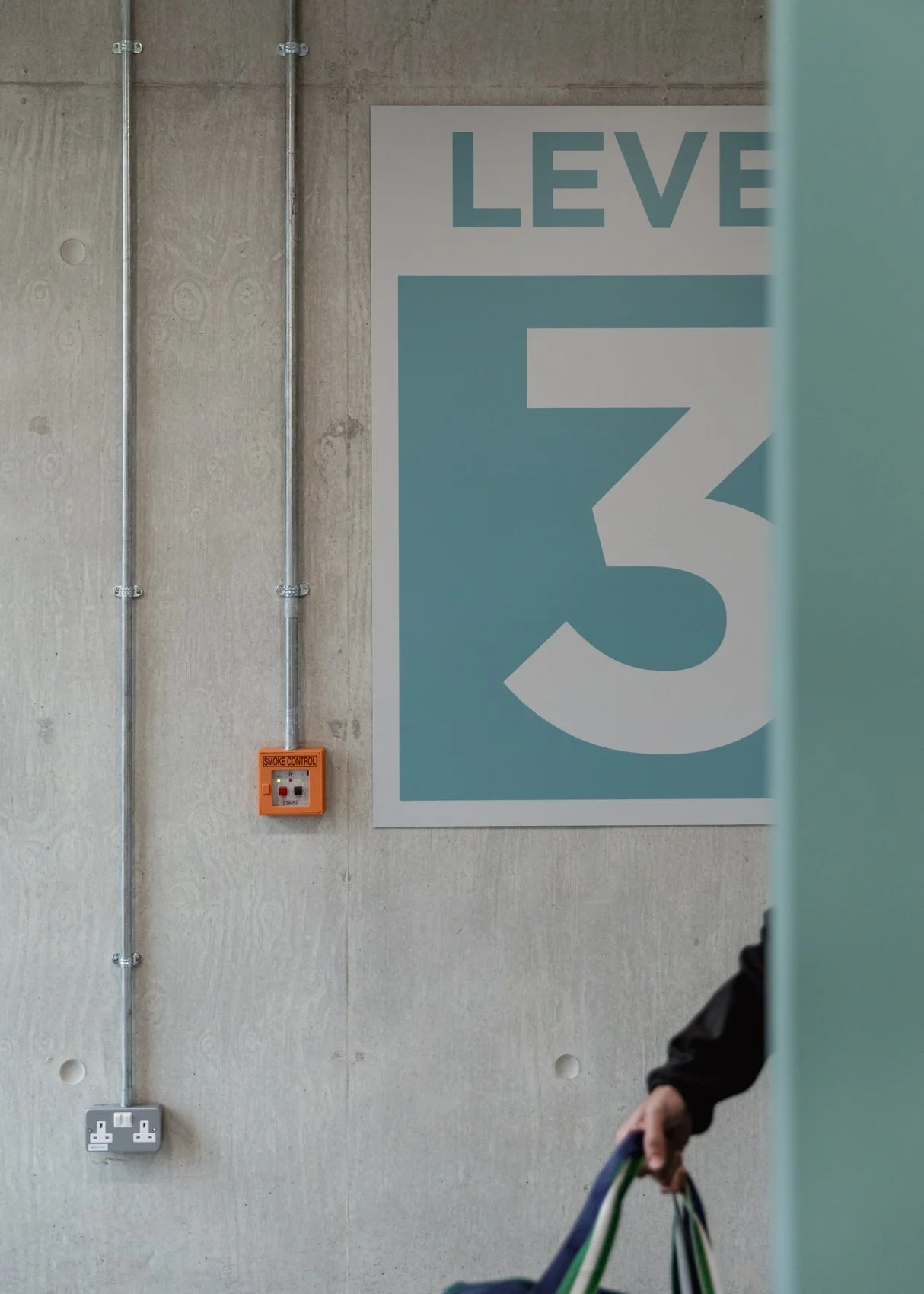 Part of a large white and teal sign displaying the number 3 and the word "LEVEL" in a parking garage or similar setting. A person's hand holding multicolored straps is partially visible in the lower right corner, and there are exposed electrical cond