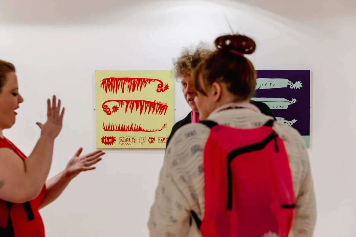 Three people in an art gallery observing colorful artwork on a white wall.