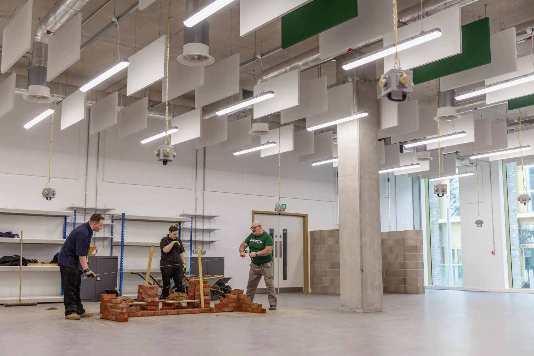 Three men building a brick wall in an industrial-style room with high ceilings, exposed ductwork, and large windows.