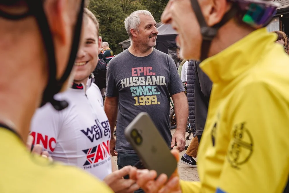 A group of smiling people outdoors, including a man wearing a dark t-shirt that says, 'Epic Husband Since 1999', and cyclists wearing helmets and colorful jackets. One person is holding a phone.