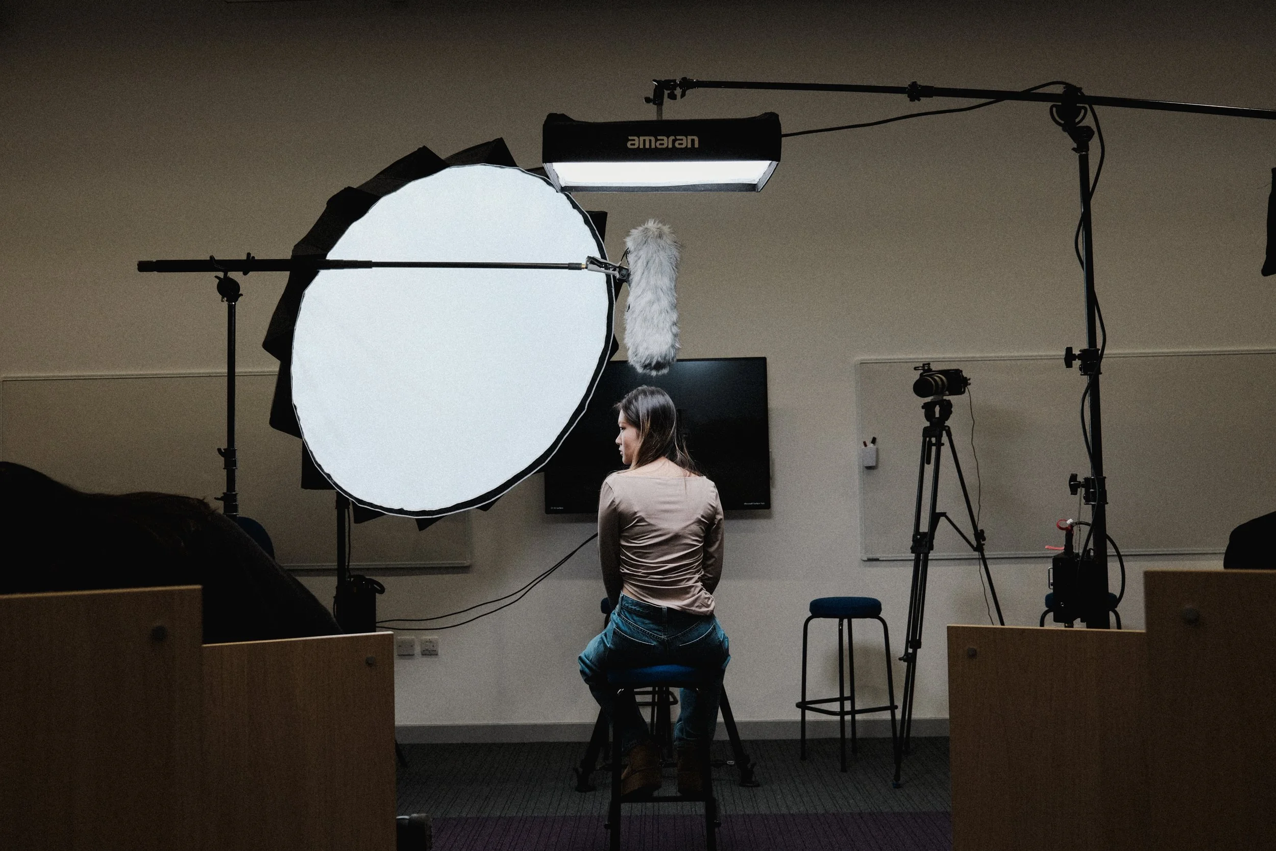 A woman sitting on a stool in a recording studio with professional lighting and camera equipment, including a large softbox and a microphone.