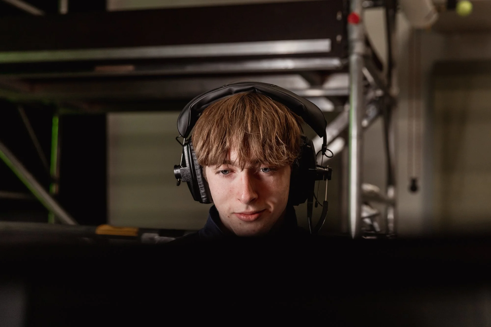 A young man with reddish-brown hair wearing a black headset, looking intently at a computer screen.