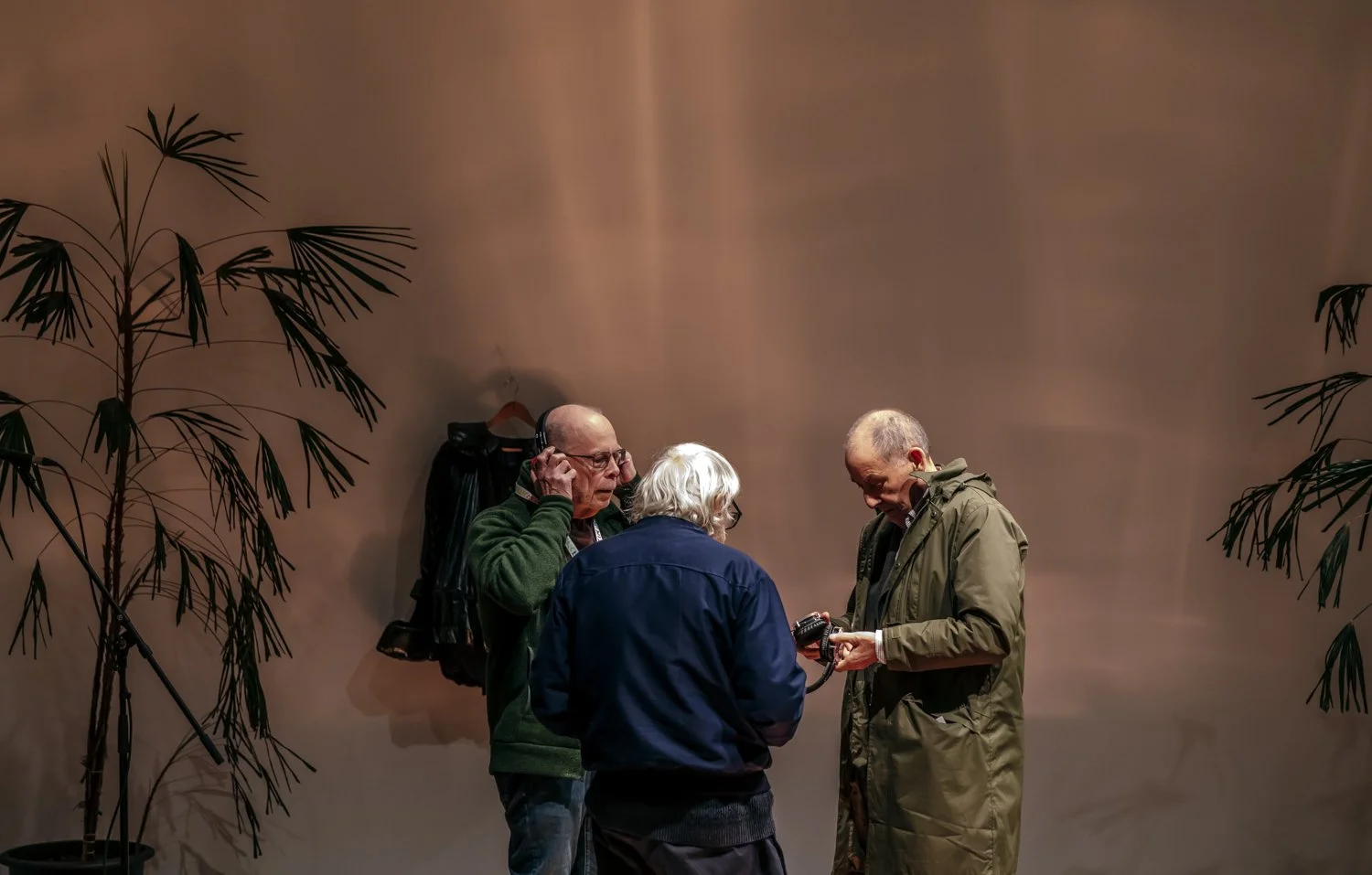 Three older men are standing together in front of a plain wall, examining a small object one of them is holding, while the third man wears headphones. There are two tall plants on either side of them.