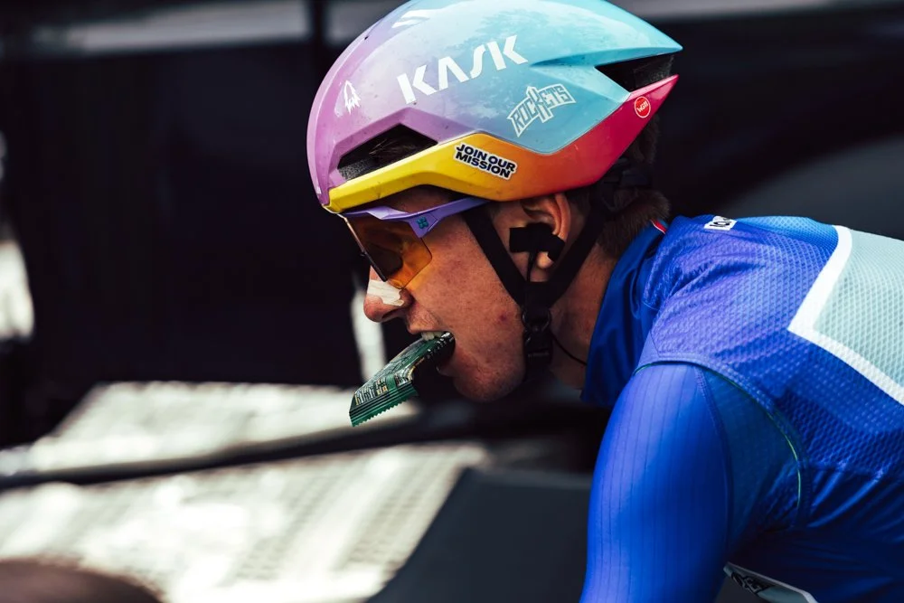 A cyclist wearing a colorful helmet, sunglasses, and a blue racing jersey, biting into a snack bar.