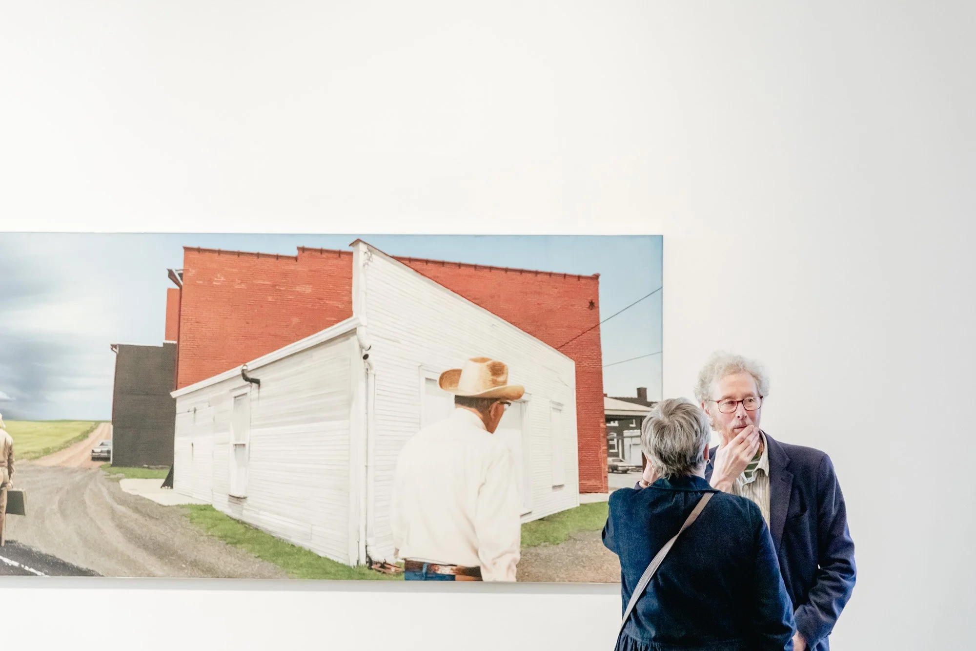 People in an art gallery observing a painting of a rural scene with a white house and a red brick building.