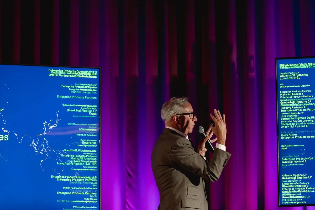 A man with gray hair and glasses, dressed in a suit, speaking into a microphone on stage with purple curtains, gesturing with his right hand, two digital screens displaying text behind him.