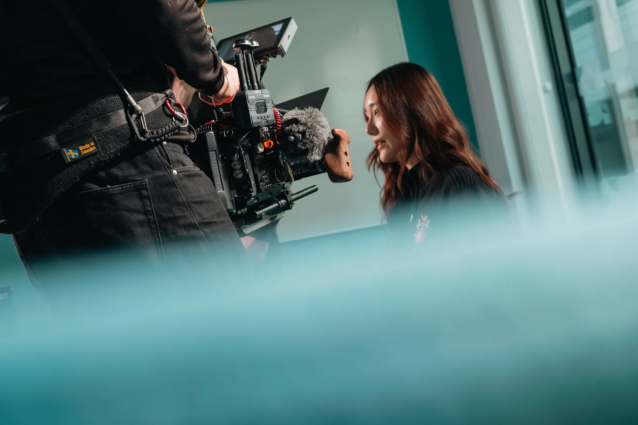 A woman with long brown hair is sitting indoors near a window, being filmed by a person with a camera.