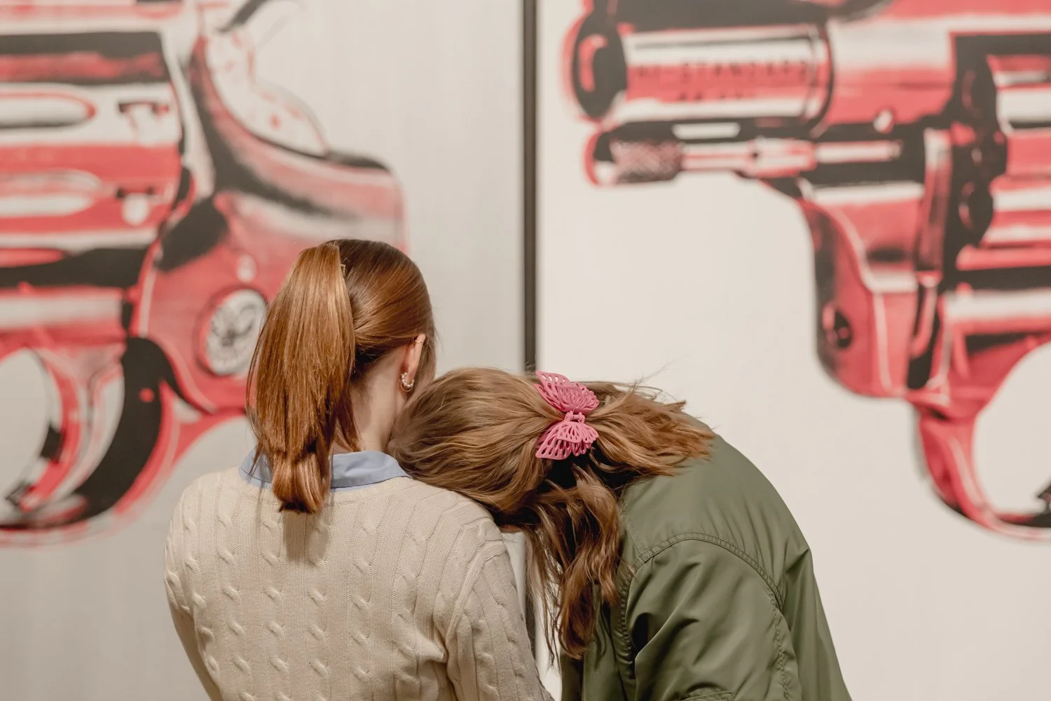 Two women with long hair, one with a pink butterfly hair clip, stand close together in front of artwork of guns at an art gallery.