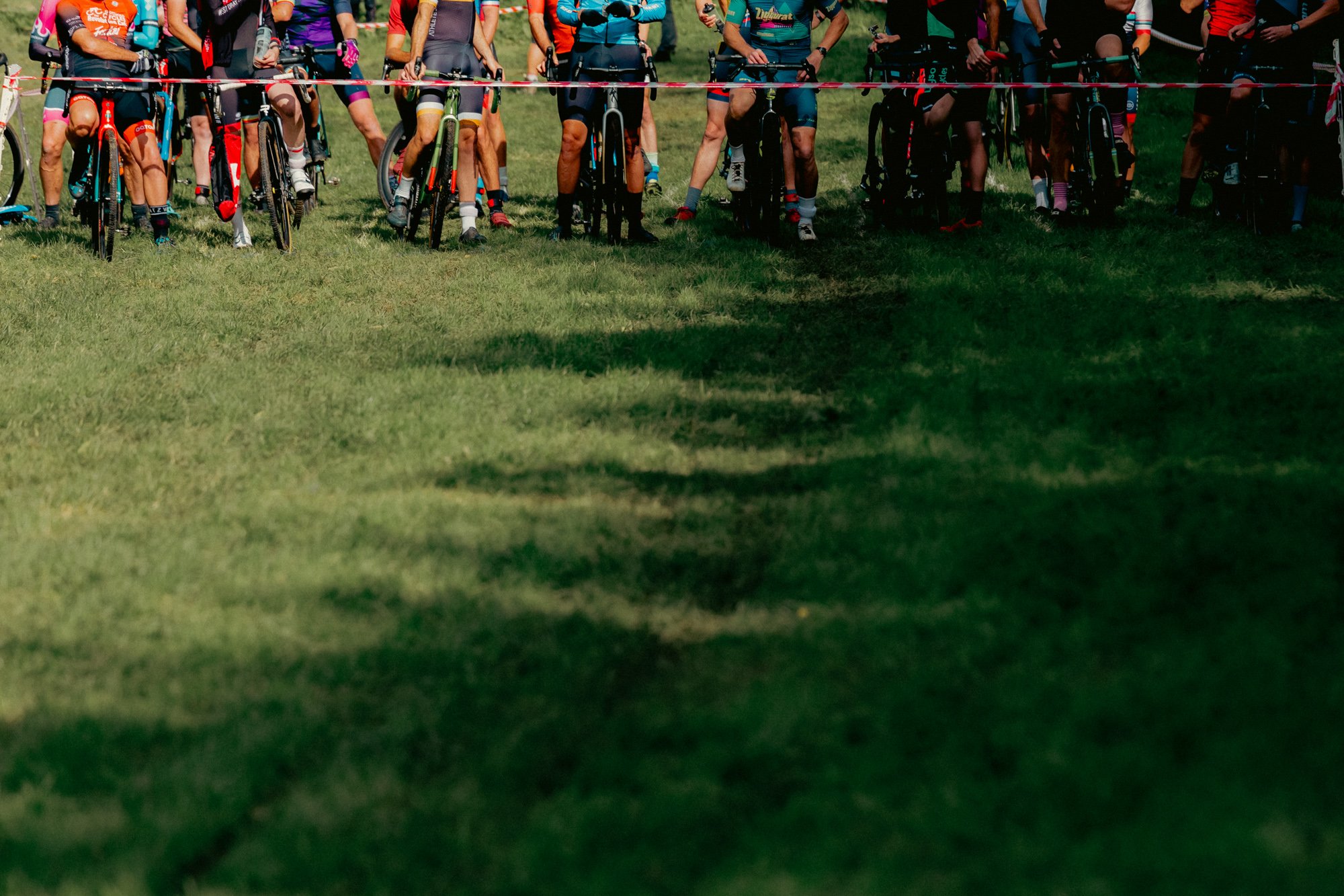Start line of a mountain biking race with cyclists on their bikes, all wearing colorful gear, standing on green grass under clear weather.