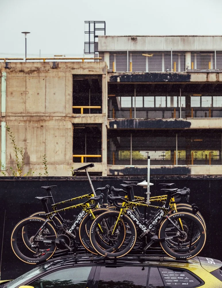 Multiple black and yellow Cervélo bikes mounted on the roof of a car, parked in front of an unfinished concrete building.