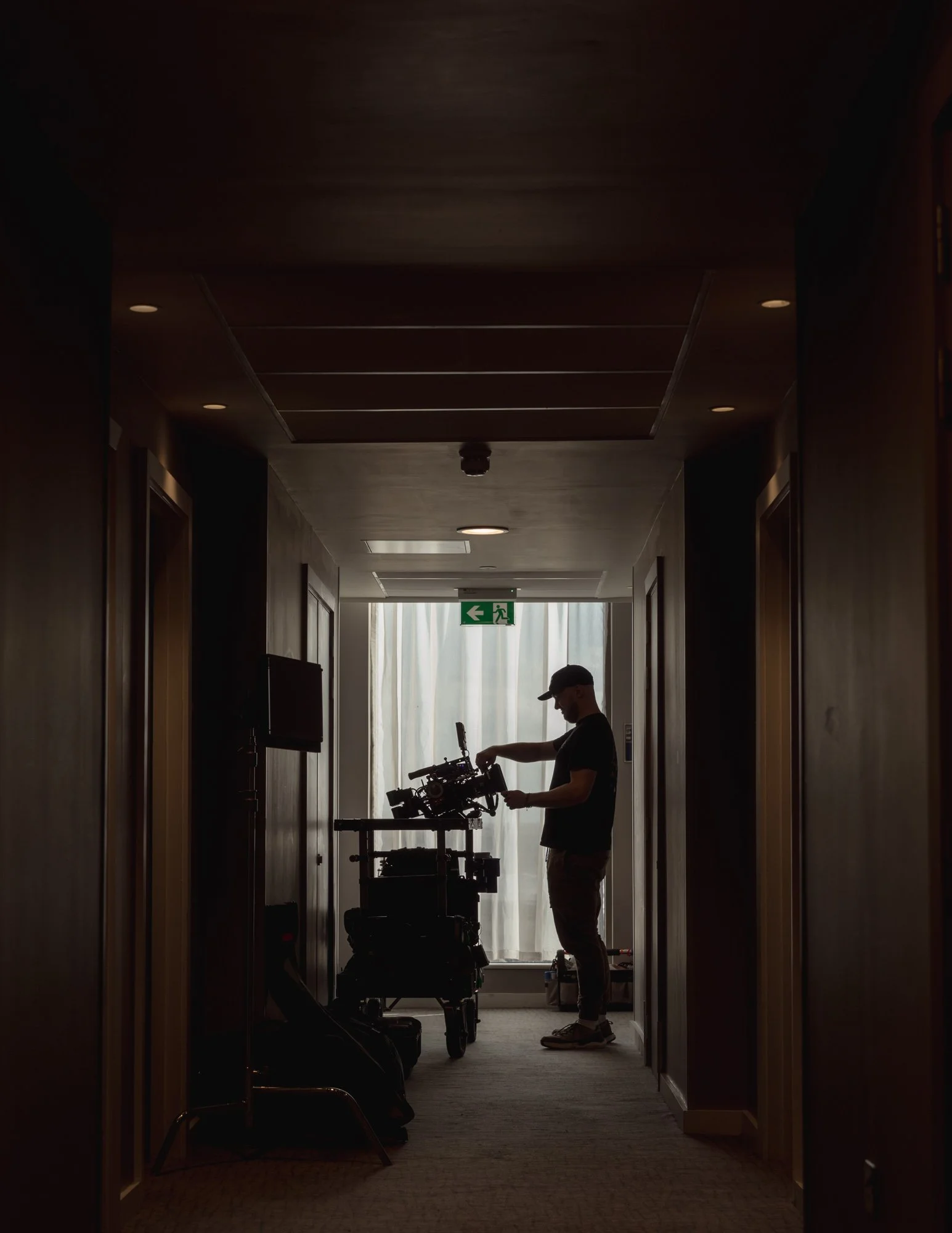 Silhouette of a person adjusting a film camera on a dolly in a dimly lit corridor with closed doors, a curtain, and an exit sign in the background.
