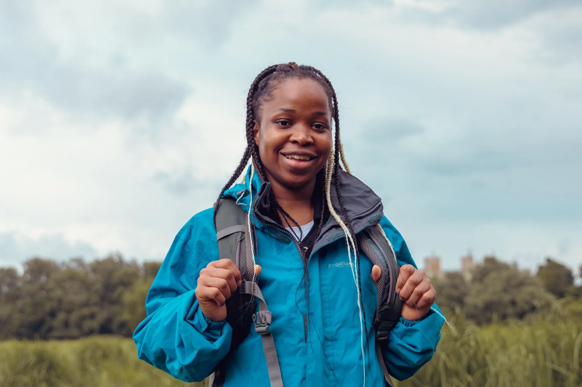 A smiling young woman with braids wearing a blue outdoor jacket and carrying a backpack, standing in a grassy field with trees and buildings in the background under a cloudy sky.