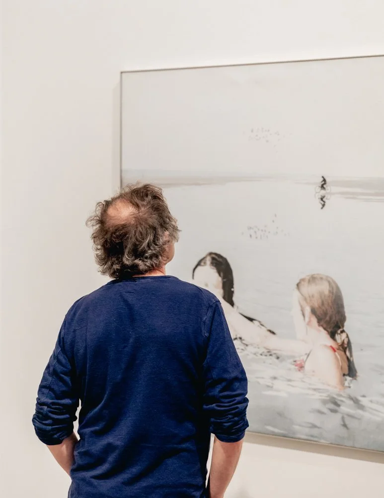 A man with curly hair and a blue shirt looking at a black-and-white photograph of two women in water.