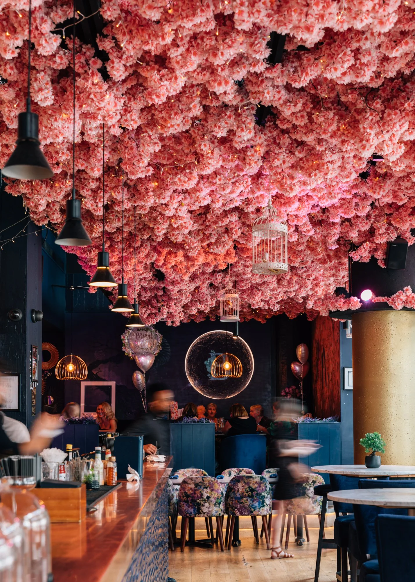 A restaurant interior decorated with a ceiling covered in pink flowers, black pendant lights, and birdcages. Customers are seated at tables and the bar.