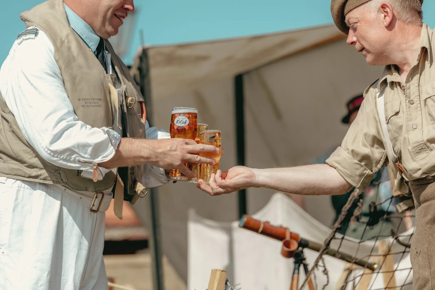 Two men exchanging drinks at an outdoor event, one man is passing a glass of beer to the other.