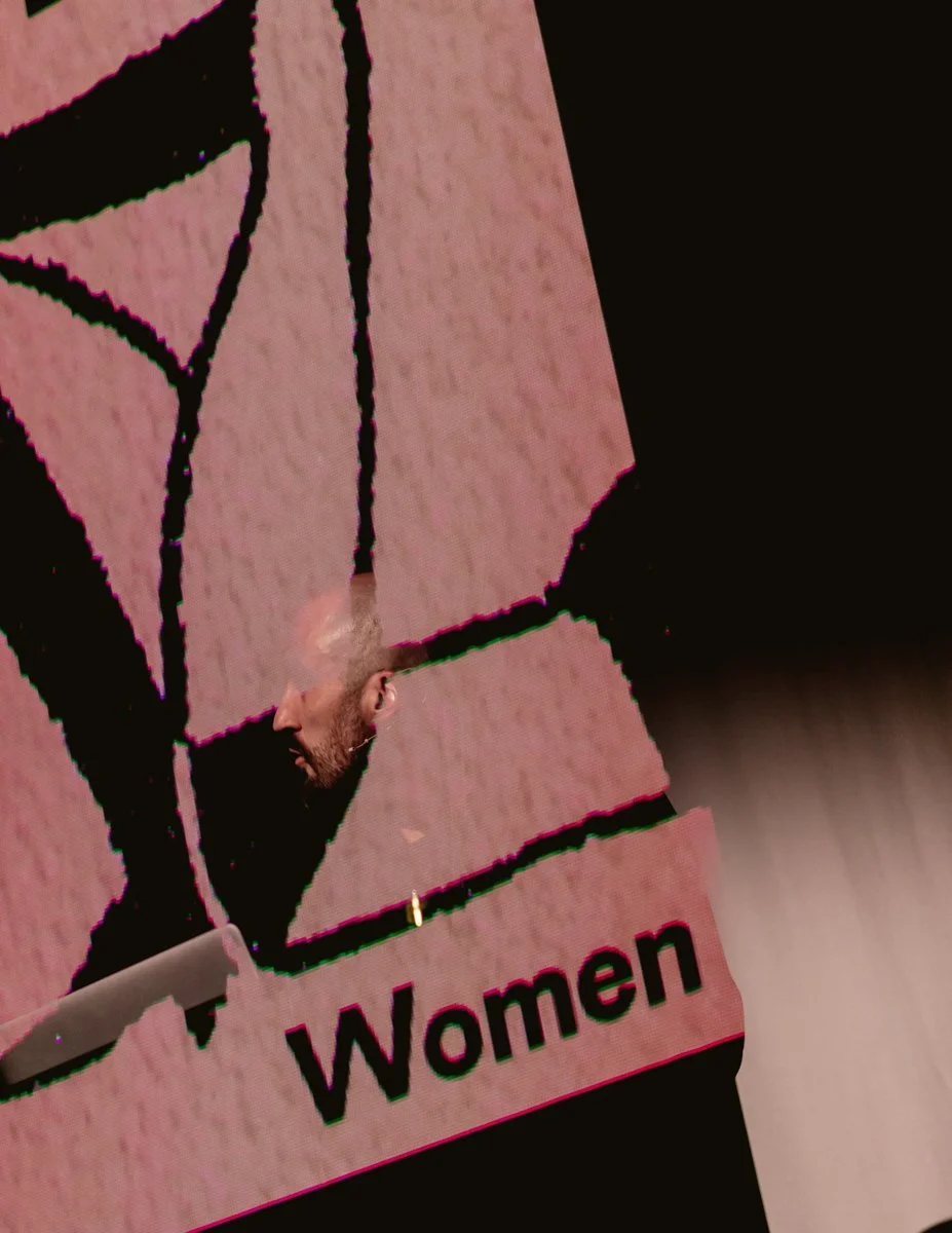Digital screen displaying a top-down view of a selfie taken in front of a women’s restroom sign. The sign features a black outline of a woman figure on a pink background with the word "Women" written below.