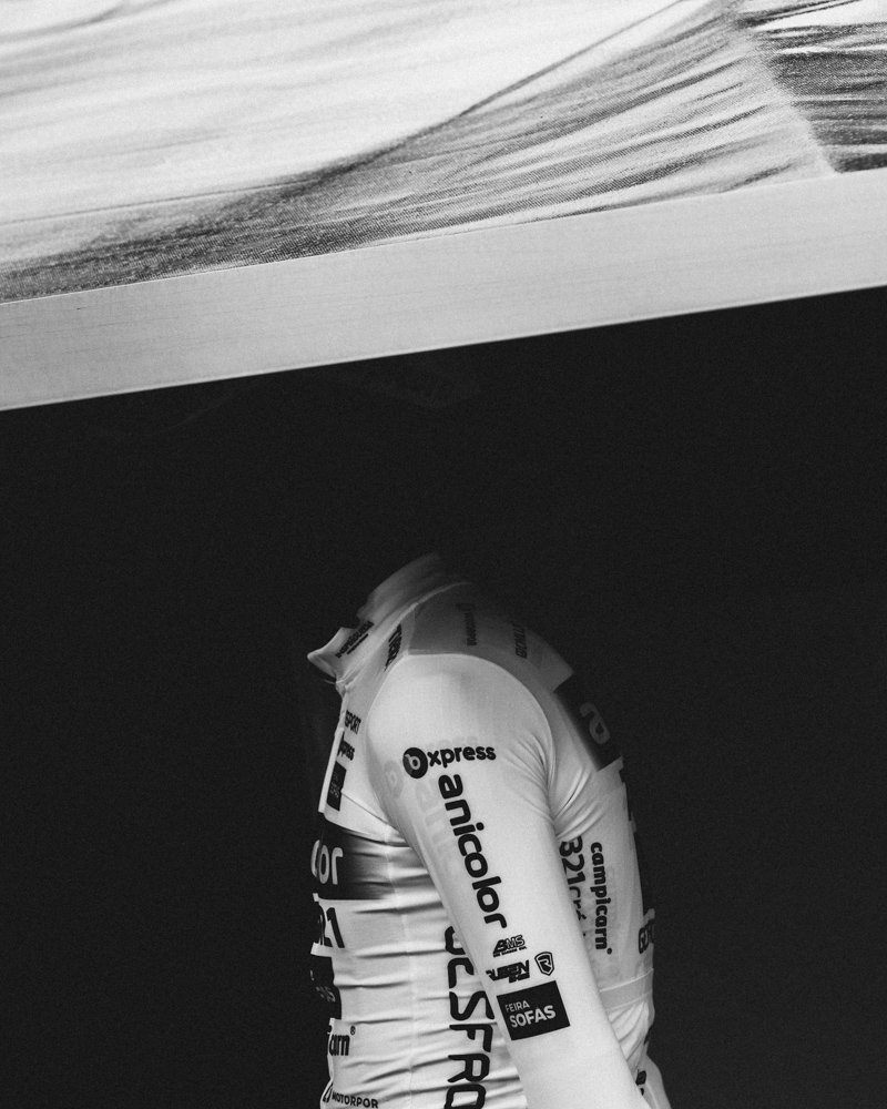A headless cyclist in a white racing jersey looking upwards, with the head cut off, under a dark ceiling, with the image in black and white.